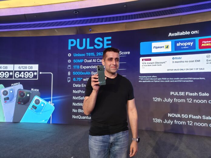 A man on stage holding up a green PULSE smartphone, with a large screen behind him displaying the phone's features like "PULSE," "Unisoc T615," "50MP Dual AI Camera," "5000mAh Battery," and "Available on: Flipkart, Shopsy." To the left, several PULSE phones in different colors (Purple, Black, Blue) are shown with pricing.