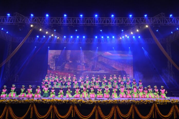 Spectacular Group Cultural Dance Performance on Stage Large group of young school girls in matching pink and green traditional costumes kneeling on stage under blue spotlights during a dance performance.